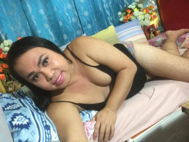xAsianSexSymbolx with black hair, pink lipstick, and a black tank top, lying on a bed, showing her tits and a slight smile, legs crossed, in a cozy room.