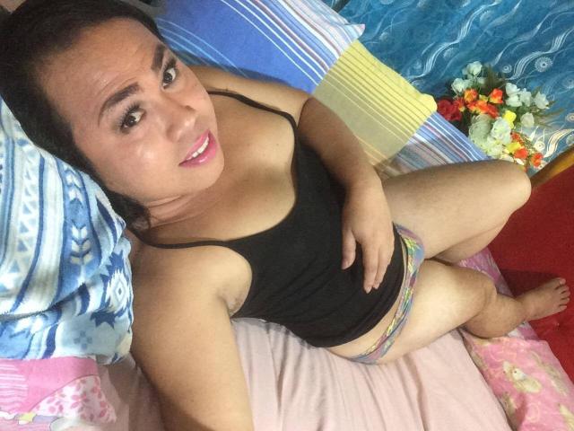 xAsianSexSymbolx in a black tank top, sitting on a bed with pink sheets, smiling at the camera, hand on her belly, colorful underwear, brown hair, and pink lipstick.
