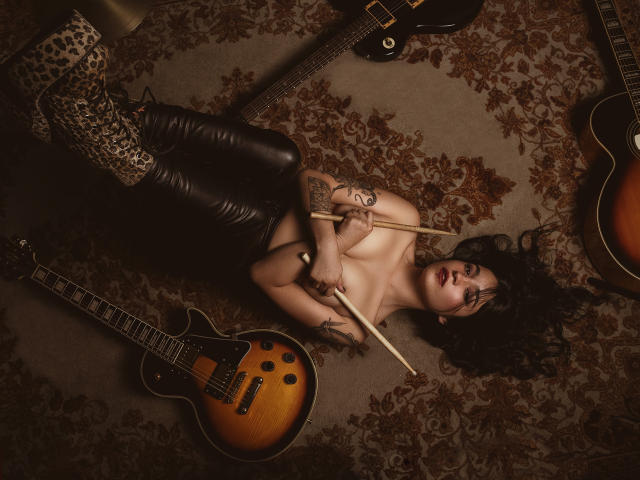 DulceHarper is lying topless on a rug, holding drumsticks, wearing leopard print boots and black leather pants, surrounded by guitars, with tattoos on her arms and side.