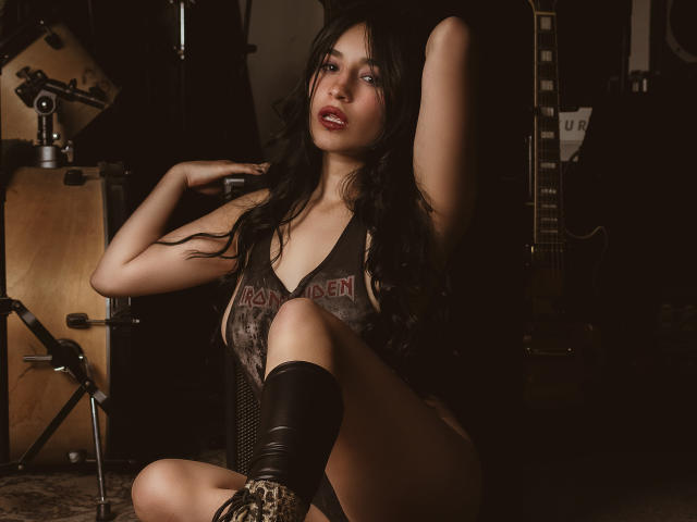 DulceHarper with long black hair, red lipstick, wearing a dark, cut-out, leather-like top, and black knee boots, sitting with one arm raised, showing her tits and ass.