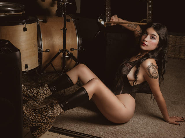 DulceHarper with leopard boots, black lace lingerie, and tattoos, sitting on carpet with legs spread, drum set in background, looking at camera with seductive expression, pale skin.