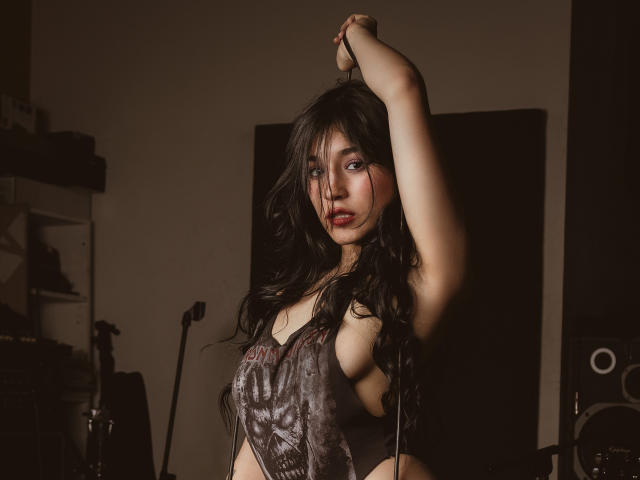DulceHarper with long black hair, wearing a revealing gray top, arm raised, showing sideboob, and a seductive look, in a dimly lit room, with a microphone behind.