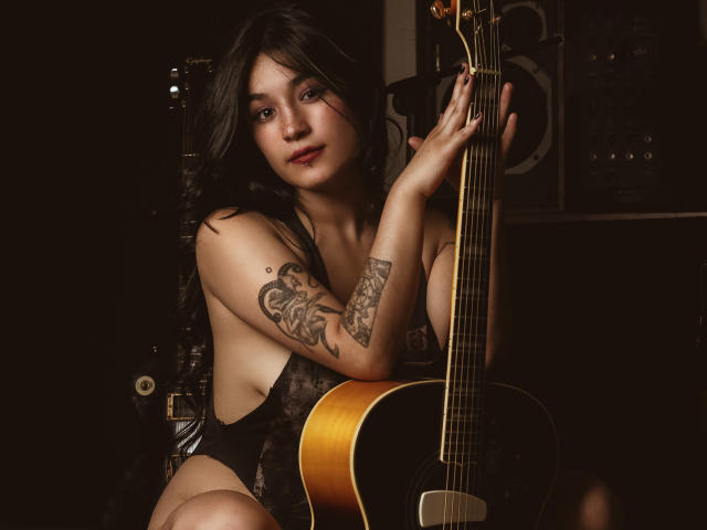 DulceHarper with dark hair, tattoos on her arm, wearing black lace lingerie, sits with a guitar, her hand on the neck, showing her tits, in dim lighting.