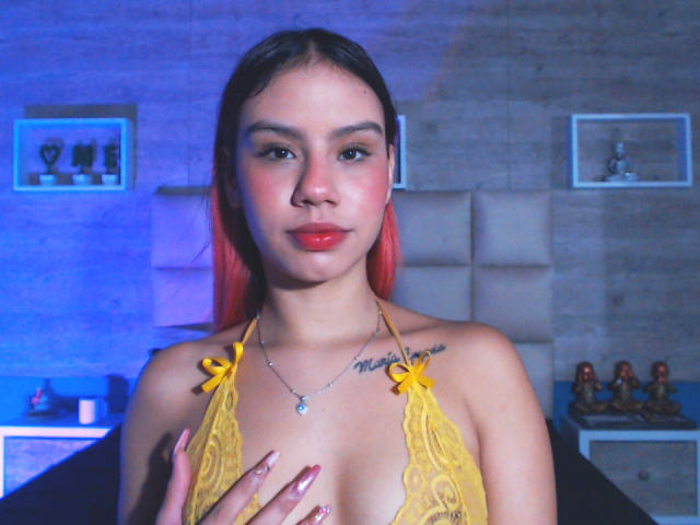 SussyGray with red-tipped hair, yellow lace bra, covering her tits with one hand, red lipstick, tattoo above right breast, in a dimly lit room, blue light.