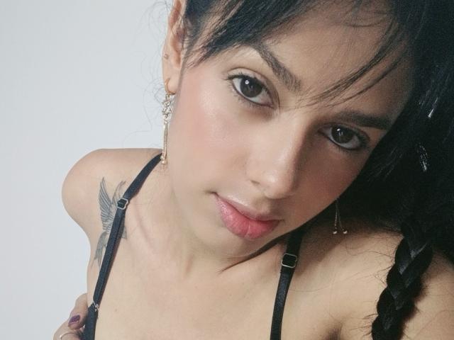 AnaisHilton has a cute face with dark eyes, pink lips, and a small nose. She has black hair in a braid, a tattoo on her right shoulder, and wears a black strap top.