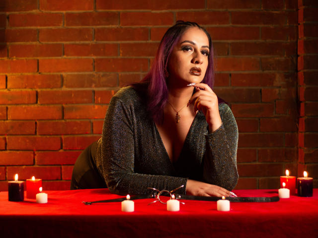 AndreaSharick with purple hair, sparkling gray top, red table, candles, glasses, and black whip, finger to lips, looking seductive. Boobs visible, dark lipstick, brick wall background.