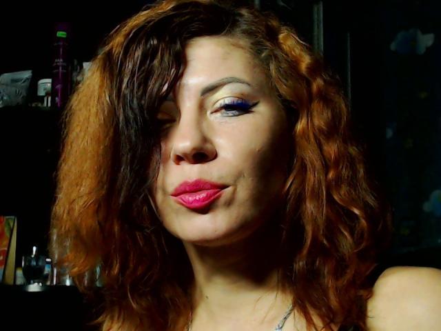 JuliaIce has wavy, half-orange half-brown hair, bold purple eyeshadow, and pink lipstick. She's winking with a slight smirk, showing her nose and lips. Dark background.
