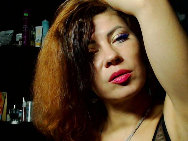 JuliaIce with wavy brown-orange hair, one eye closed, pink lipstick, purple eyeshadow, arm raised, black mesh top, looking seductive. Shiny skin, dark background.