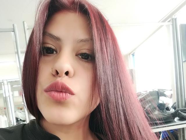 ElaiaaBakerr has red hair, a nose ring, and pink lipstick with puffy, glossy lips. She's wearing a black shirt, and her eyes are slightly open.