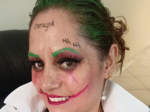 NatashaChaus with green eyeshadow, pink and red makeup on face, green hair, white collar, and "HA HA HA" written on forehead. Her tits are visible in the lower part.