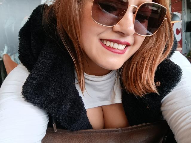 NatashaChaus with brown hair, red lipstick, and large tits, wearing a white shirt, black fuzzy vest, and gold sunglasses, smiling at the camera. Her boobs are slightly visible.