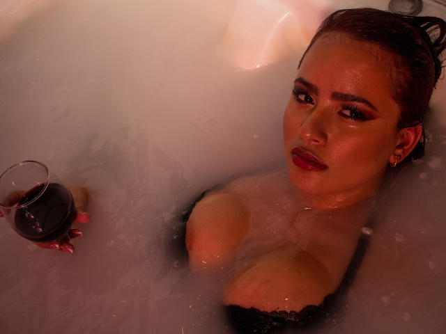 LaurenMartins with wet brown hair, red lipstick, and black lace top, holding a glass of red wine, in a white bathtub, looking at the camera with a seductive smile.