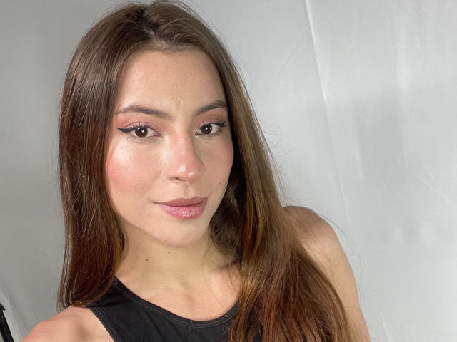 DaphneBlakee has long brown hair, pink eyeshadow, and a slight smile. She's wearing a black top, with a shiny, slightly flushed face. Her tits are small and cute.