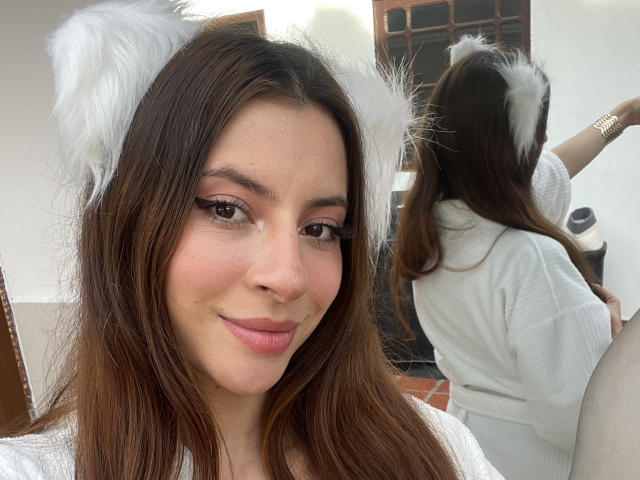 DaphneBlakee with white furry cat ears, brown eyes, and pink lipstick, smiling at the camera, reflection showing her back in a white top, brown hair, and gold bracelet.