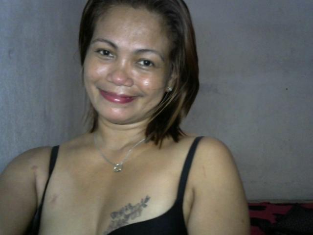 CuteDonna with short brown hair, smiling, wearing a black bra, showing small tits with a tattoo above her chest, wearing a necklace, has light skin and red lipstick.