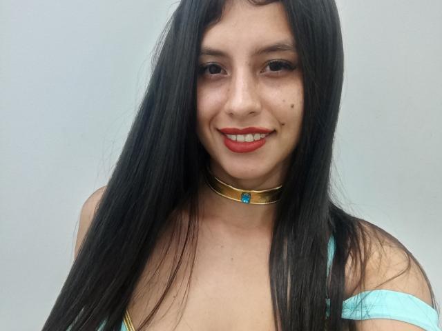 MadissonFontaine with long black hair, red lipstick, and a gold choker with a blue gem, smiling at the camera, wearing a teal top, showing small tits.