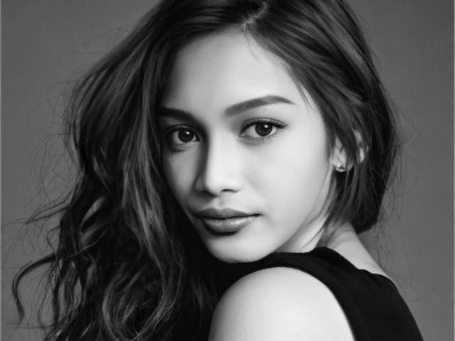 PrincessJasmina's face, gray-scale, with long wavy hair, subtle makeup, and a slight smile, looking over her shoulder, showing her bare shoulder and black sleeveless top.