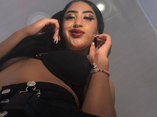 MichellDiaz, con la pelle scura, i lunghi capelli neri e il rossetto rosso, indossa un reggiseno nero e pantaloni neri abbottonati, mostrando il seno e guardando in basso con un sorriso sicuro..