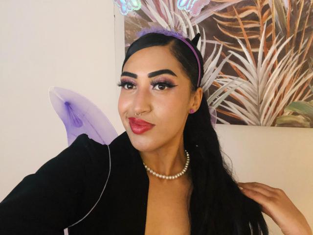 MichellDiaz, com cabelo escuro, uma faixa de cabeça roxa, um colar de pérolas, uma blusa preta, batom vermelho, um piercing no septo, delineador roxo e um sorriso divertido, segurando um leque roxo..