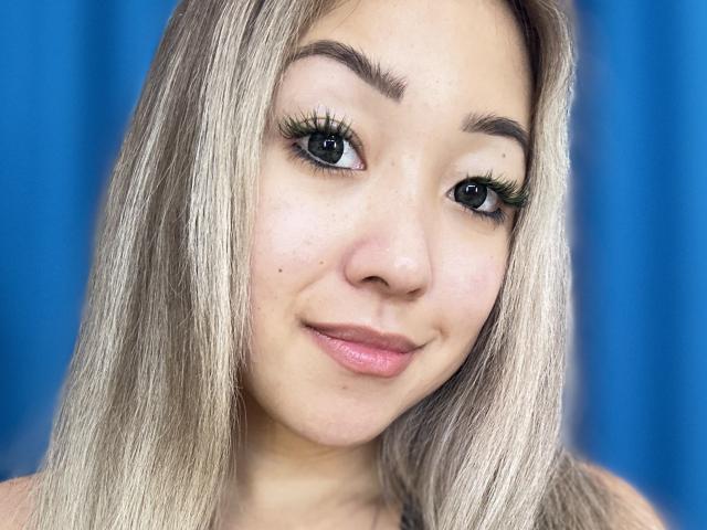 RenyLime has long, straight blonde hair, thick black eyelashes, and pink lips. She's smiling slightly with a small beauty mark on her left cheek. Her tits are not visible.