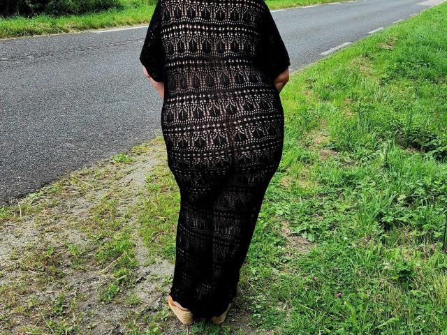 Enchaleur69X's black, crocheted dress shows a patterned, see-through butt and thick, curvy ass standing on grass by a road, wearing beige shoes.