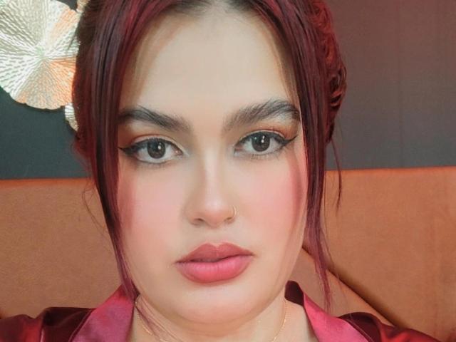 AbbyLester has red hair, dark eyebrows, eyeliner, and pink lipstick. She's looking at the camera with a neutral expression, wearing a pink satin shirt.