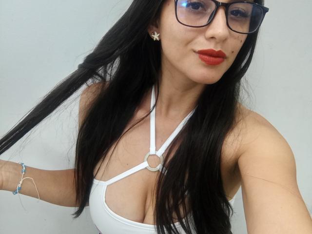 MadissonFontaine with long black hair, wearing glasses, white strappy top, red lipstick, holding hair, showing her tits, bracelet on left wrist, star earrings, smiling.