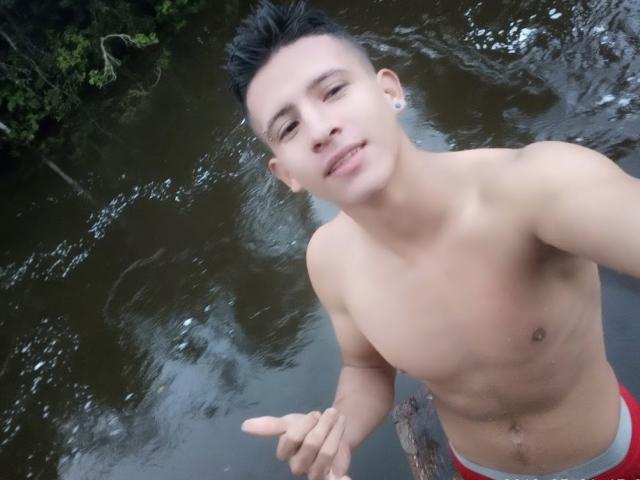 FireLion, shirtless, with short black hair, small ear piercings, and a slight smile, pointing with right hand, wearing red and white underwear, standing in a river.