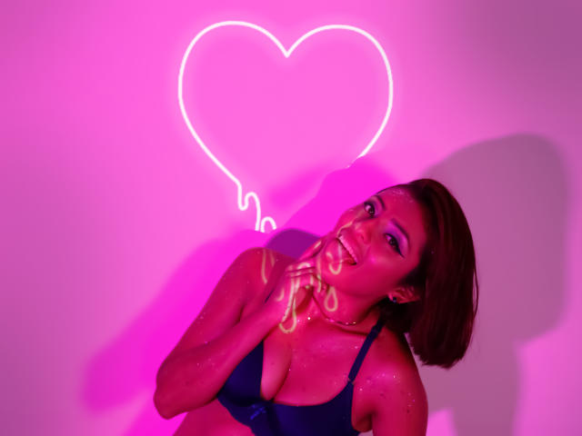 LizBonnet with short brown hair, wearing a blue bra, has pink neon heart glow behind her, smiling with a hand near her mouth, pink light highlighting her shiny skin.