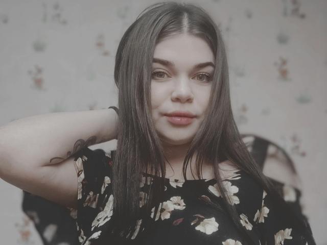 SophieApple with long brown hair, floral black dress, hand behind head, subtle smile, soft makeup, fair skin, small tattoo on upper arm, slightly blurred background, gentle light.