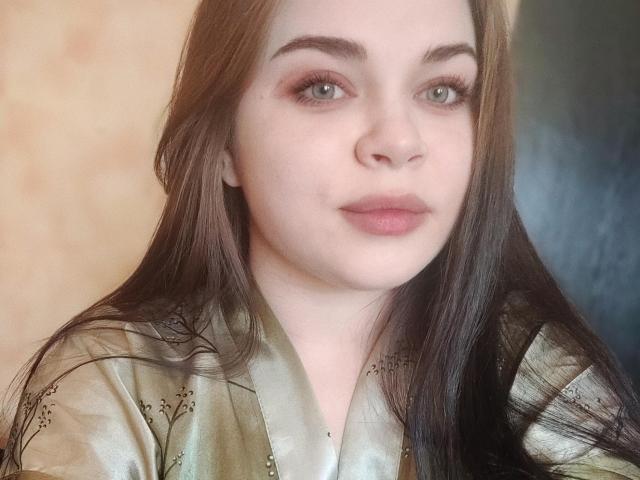 SophieApple with long brown hair, green eyes, and pink lips, wearing a shiny green silk robe with subtle floral patterns, looking slightly to the right.