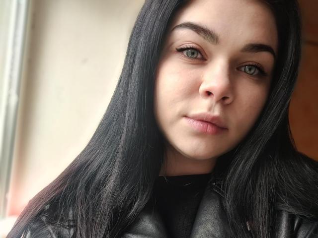 SophieApple with long black hair, green eyes, and a slight smile, wearing a black leather jacket, looking directly at the camera. Her face is slightly tilted to the right.