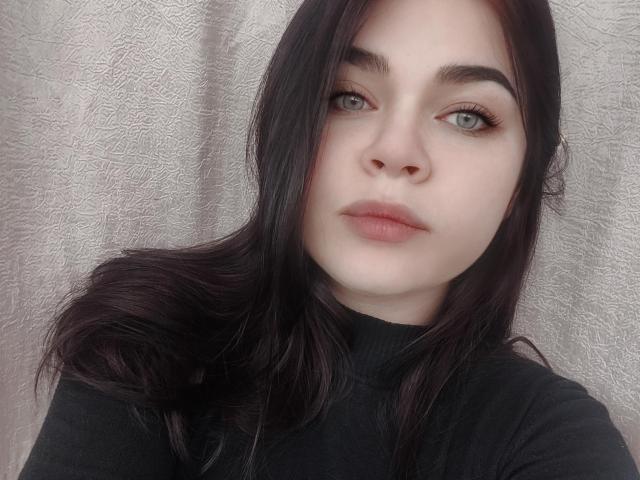 SophieApple has pale skin, dark brown hair, and blue eyes. She's wearing a black turtleneck, with her lips slightly parted and a neutral expression on her face.