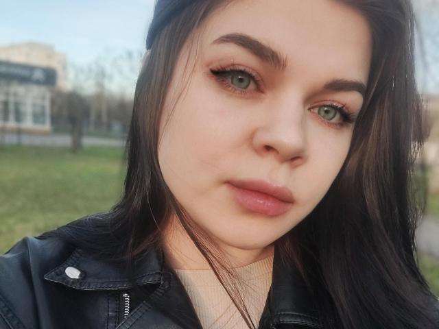 SophieApple has green eyes, full pink lips, and long dark brown hair, wearing a black jacket, with a slightly tilted head and a neutral expression.