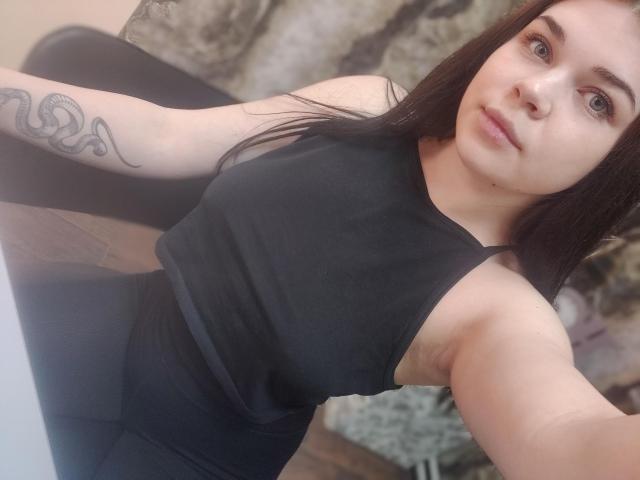 SophieApple with long black hair, wearing a black sleeveless top and black pants, showing a snake tattoo on her right arm, looking at the camera with a neutral expression.
