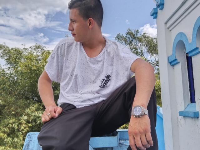 FireLion is a dude with short black hair, wearing a white dotted t-shirt, black pants, and a silver watch, sitting on blue steps, looking to the side.