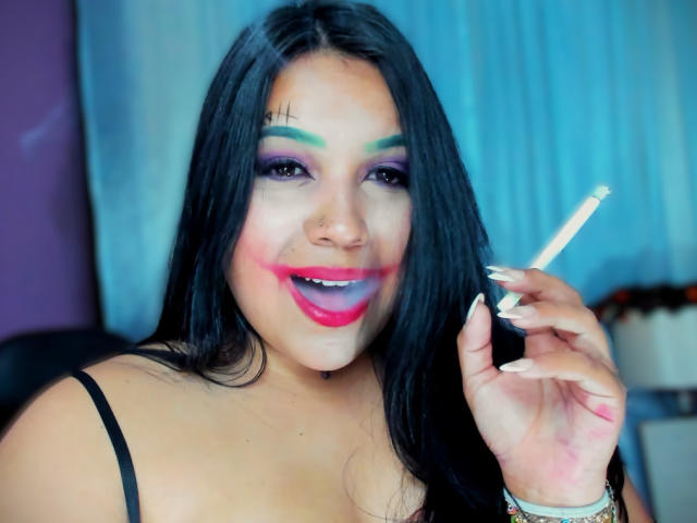 AvaChanne with long black hair, green and purple eye makeup, pink lipstick, holding a lit cigarette, smiling, nose pierced, black strap, blue and purple background, painted green eyebrows.