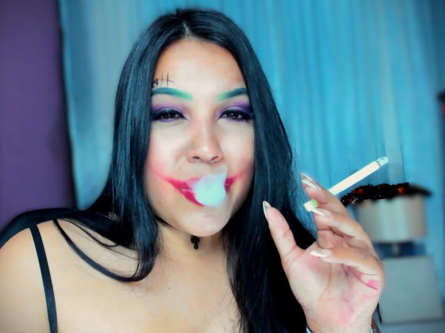 AvaChanne with long black hair, colorful eye makeup, and pink lipstick smeared with white cream, holding a lit cigarette, wearing a black strap top, showing her tits.