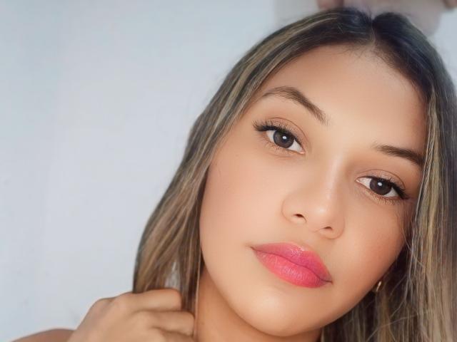 SashaChloe has light brown skin, long brown hair, thick eyebrows, long eyelashes, pink lipstick, and a cute face. She's touching her hair with her right hand.