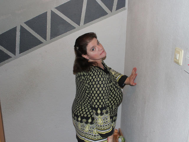 MollyBoobs-hot with large tits, wearing a green and black patterned dress, standing against a white wall, looking back with a slight smile, right hand on the wall.