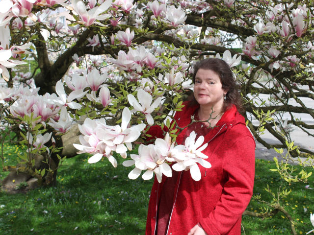 MollyBoobs-hot stands in a red coat, holding pink and white magnolia flowers, with a green grassy background and a blooming tree behind her.