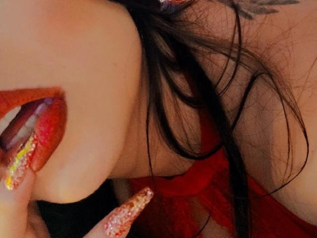 Sharp shot of Sharonscarlet's face showing bright red lipstick, glossy red nails with glitter, and black hair, with a tattoo on the right side of her neck.