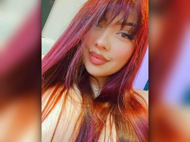 Sharonscarlet has red and purple dyed hair, a cute smile, and full lips with a slight pout. Her makeup is subtle, and she's wearing a white top.