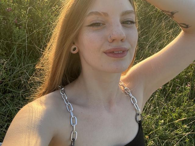 ElsaBj with light brown hair, nose ring, and septum piercing, wearing a black top with silver chain straps, smiling, arms raised, sunlit, grassy background, tattoo on right arm.