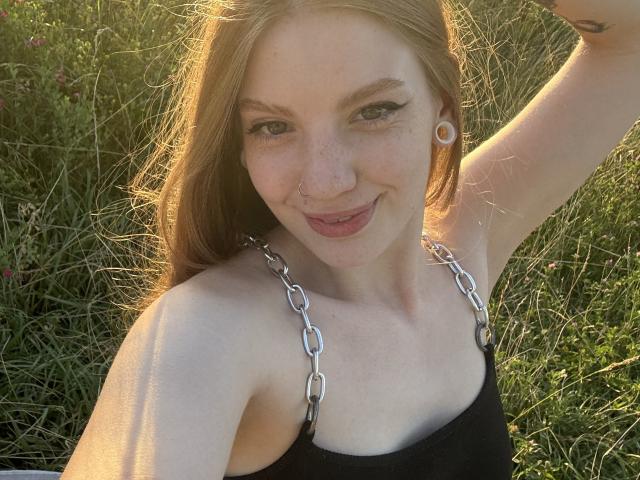 ElsaBj with light brown hair, nose ring, and white ear plugs, smiling at camera in black tank top with silver chain straps, arm raised, sunlight on face.