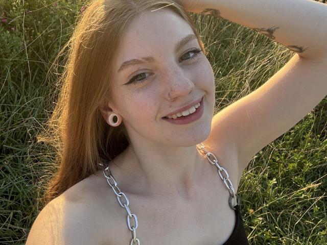 ElsaBj has pale skin, red hair, green eyes, and a nose ring. She's smiling with her arm raised, showing a tattoo, wearing a black top with silver chain straps.