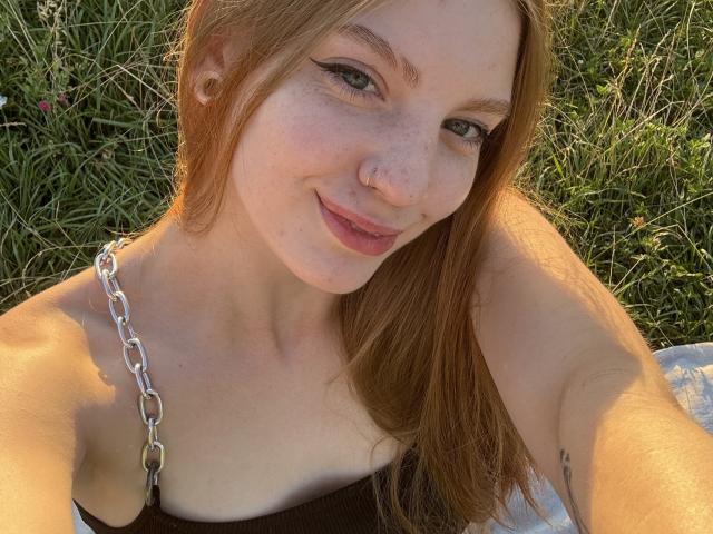 ElsaBj with freckles, green eyes, and nose ring, smiling, red hair, wearing a brown top with a chain strap, outdoors, selfie, showing her tits, tattoo on left arm.