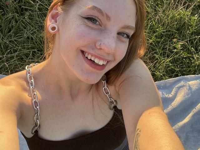 ElsaBj with a big smile, freckled face, nose ring, white ear gauges, chain necklace, brown tank top, and a tattoo on her right arm, lying on grass.
