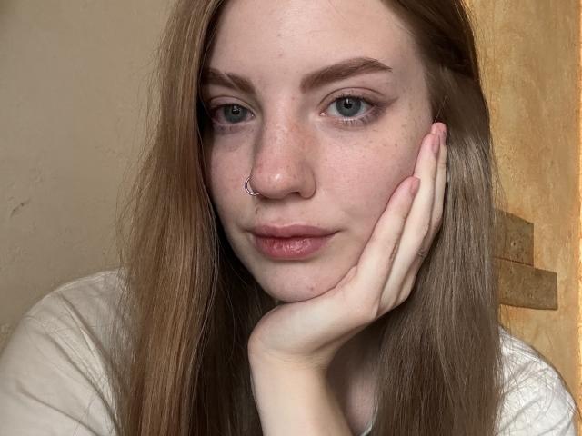 ElsaBj has light green eyes, a nose ring, and freckles. She's resting her face on her hand with long brown hair, wearing a white shirt.