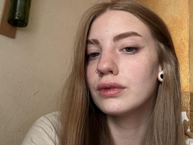 ElsaBj has pale skin, long blonde hair, green eyes, and a nose ring. She has small black ear plugs and pink lips. She's wearing a white top.